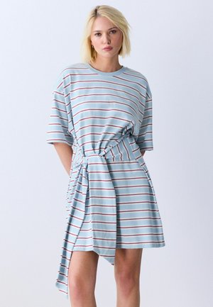 Blondynka w jasnoniebieskiej, czerwonej i białej oversize sukience w poziome paski z wiązanym detalem w talii, stojąca na jednolitym tle.