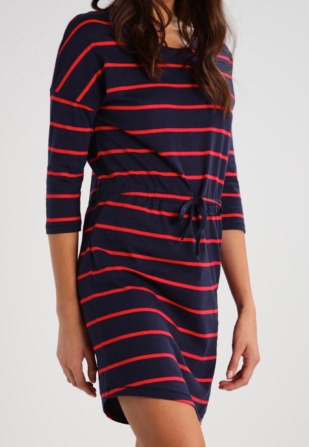 Femme portant une robe bleu marine avec des rayures horizontales rouges et une taille à cordon, debout devant un fond clair uni.