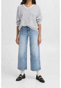 Donna che indossa un maglione a righe bianche e blu navy con scollo a V, jeans a gamba larga azzurri, calzini bianchi e mocassini neri con dettagli dorati.