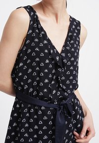 Svart ärmlös klänning med V-ringning, prydd med vita triangulära mönster. Inkluderar ett knytbart midjebälte och mjuk, texturerad tyg.
