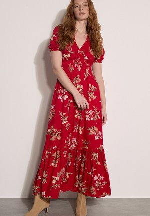 Kobieta o długich rudych włosach, ubrana w czerwoną kwiecistą sukienkę maxi z bufkami na rękawach oraz beżowe botki na kostkę, stojąca przy jasnej ścianie.