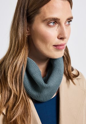 Strikket cowl i blødt, tekstureret stof, der har en lys blågrøn farve. Bæres sammen med en beige frakke og en marineblå top, hvilket fremhæver dets hyggelige design.