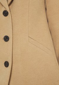 Abrigo de lana beige con un acabado texturizado, seis botones oscuros y un bolsillo lateral. La vista en primer plano captura los detalles de la tela y la construcción.