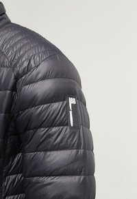 Veste matelassée noire avec détails matelassés. Comprend une poche appliquée avec un logo sur l'épaule gauche et une texture lisse et brillante.
