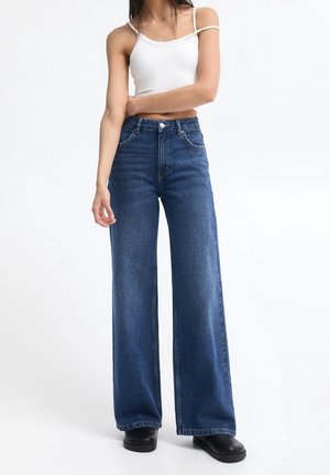 Femme portant un jean bleu taille haute à jambes larges, un crop top blanc avec fines bretelles, et des chaussures noires, debout sur un fond blanc uni.