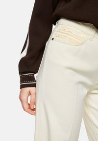 Weiße weit geschnittene Jeans mit einem glatten Material, ausgestattet mit dekorativer gelber Naht an den Taschen. Ein braun-weiß gestreiftes Langarmshirt ist teilweise sichtbar.