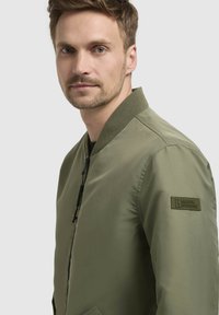 Olivgrüne Bomberjacke mit Reißverschluss vorne, geripptem Kragen und einem Patch am Ärmel. Aus glattem, leichtem Stoff mit einem minimalistischen Design.