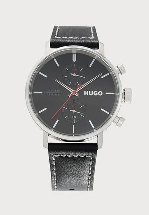 Montre-bracelet Hugo noire avec boîtier argenté, index des heures blancs, aiguille des secondes rouge et bracelet en cuir noir avec coutures blanches.