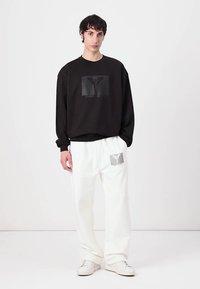 Čierna mikina s textúrovanou grafickou značkou a biele tepláky so značkovou nášivkou na nohe. Biele tenisky dopĺňajú look.