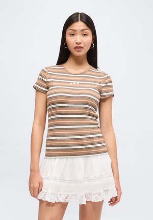 Young woman with long dark hair wearing gold hoop earrings, a brown striped fitted T-shirt, and a white ruffled skirt.