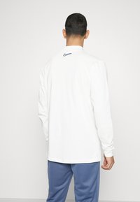 Long-sleeve white T-shirt with a smooth texture, featuring a small dark logo at the upper back. Paired with blue pants.