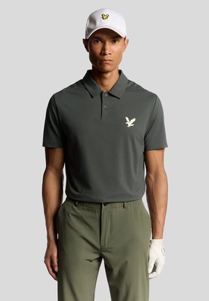 Mørkegrøn poloshirt med et diskret ørnlogo, parret med lys grønne bukser og en hvid cap med et gult ørn emblem. Hvid golfhandske.
