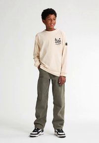 Maglietta beige a maniche lunghe con stampa grafica nera, abbinata a pantaloni verde oliva e sneakers bianche e nere. Design semplice e vestibilità rilassata.