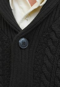 Schwarze gestrickte Strickjacke mit einem strukturierten Kabelmuster und Rippenstruktur. Besitzt einen großen grauen Knopf und ist mit einem weißen Hemd mit Kragen kombiniert.