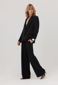Kobieta o blond włosach ubrana w czarny oversize'owy garnitur, srebrną bransoletkę na nadgarstek i wysokie obcasy w leopardzi wzór, stojąca na neutralnym tle.