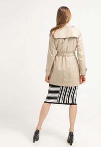 Beżowy trencz z paskiem w talii, ozdobiony guzikami i z rozcięciem z tyłu, w zestawie z czarno-białą spódnicą w paski.