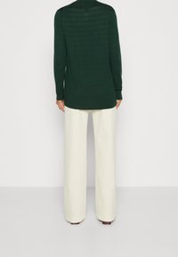 Chaqueta verde de manga larga con una ligera textura, dobladillo redondeado y puños acanalados, combinada con pantalones anchos de color crema.