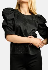 Čierna blúzka s nafúknutými rukávmi, posiata malými striebornými ozdobami, s okrúhlym výstrihom. Vyrobená z hladkej tkaniny so štruktúrovaným strihom.