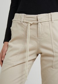 Beige cargo broek met een tailleband met trekkoord, zijzakken en een gestructureerde stof. Kenmerkt zich door stiksel details aan de voorkant.