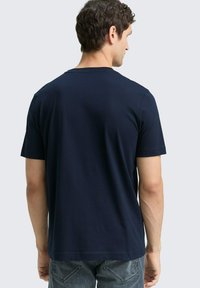 Granatowy t-shirt z krótkim rękawem wykonany z gładkiego, miękkiego materiału. Podstawowy fason z okrągłym dekoltem, bez wzorów i ozdób, widoczny od tyłu.