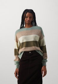 Pull en tricot avec des rayures vertes, beige, crème et olive. Texture côtelée, col rond et coupe ample. Assorti à une jupe marron foncé.