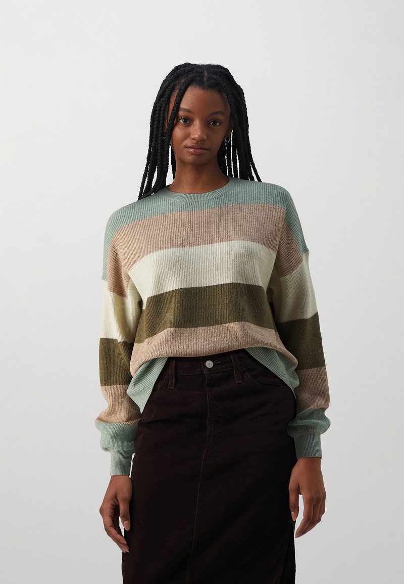 Pull en tricot avec des rayures vertes, beige, crème et olive. Texture côtelée, col rond et coupe ample. Assorti à une jupe marron foncé.