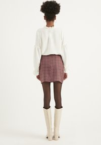 Persona sta in piedi di spalle indossando una blouse bianca con colletto a ruffle, una gonna mini a scacchi bordeaux, collant neri trasparenti e stivali bianchi al ginocchio.