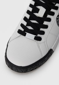 Balti odiniai sportbačiai su tekstūruotu paviršiumi, juodom raišteliais ir juodu guma padu, su įspaustu logotipo detaliu šone.