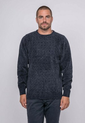 Homme aux cheveux courts et à la barbe portant un pull en maille texturée bleu foncé et un pantalon sombre, debout devant un fond clair uni.