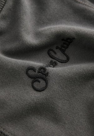 Tissu gris au toucher lisse avec un texte noir brodé indiquant "Sea's Club", mettant en avant un design simple et minimaliste.