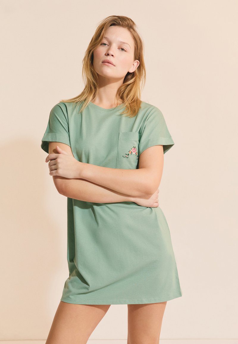 Młoda kobieta stoi ze skrzyżowanymi ramionami, ubrana w jasnobeżową, oversize'ową sukienkę t-shirt z małą kwiatową wyszywanką na kieszeni.