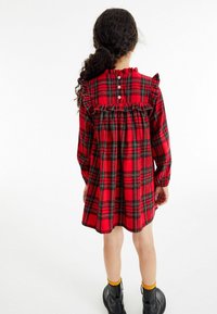 Vestido de cuadros rojos con escote con volantes y mangas largas. Presenta botones en la parte posterior y un dobladillo acampanado, combinado con botas negras y calcetines amarillos.