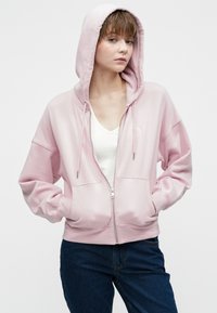 Young person with short hair wearing a light pink zip-up hoodie over a white top and blue jeans, posing with hands in pockets against a plain background.