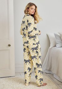 Nainen seisoo paljain jaloin keltaisissa ruudullisissa pyjamissa, joissa on mustavalkoisia seepraprinttejä, katsellen olkansa yli kirkkaassa makuuhuoneessa.