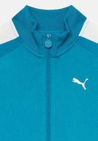 Tealfärgad sportjacka med ribbad krage och dragkedjestängning. Har en vit Puma-logga och kontrasterande vita axelpartier. Slät textur.