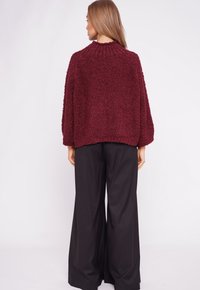 Burgundy textured sweater with a boxy design, round neck, and long sleeves, paired with black wide-leg trousers. Smooth fabric finish.