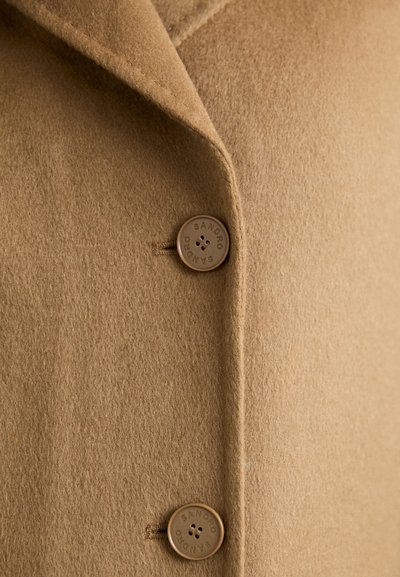 Beige uldblandingsfrakke med indsnitkrave, to brune knapper med præget brandnavn, og blødt, struktureret stof.