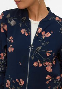 Kobieta ubrana w granatową kurtkę z różowymi kwiatowymi nadrukami i srebrnym suwakiem, nałożoną na biały top.