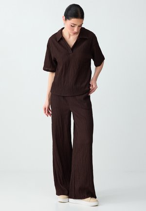 Kobieta w luźnej, ciemnobrązowej, teksturowanej koszuli z krótkim rękawem i spodniach o szerokich nogawkach, patrzy w dół z ręką przy biodrze, na białym tle.