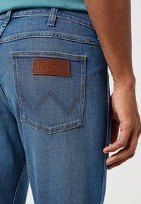 Jeans in denim blu presentano un'etichetta in pelle marrone con il testo "Wrangler" sopra un design di tasca ricamato distintivo. Tessuto strutturato visibile.