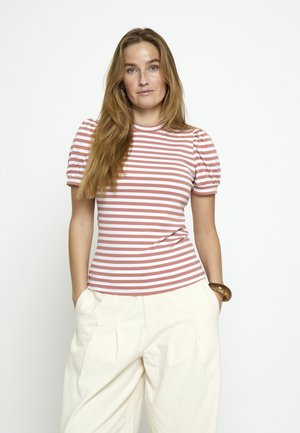 Ženska z dolgimi blond lasmi, oblečena v roza in belo črtasto kratko majico ter široke bele hlače, stoji z rokami v žepih.