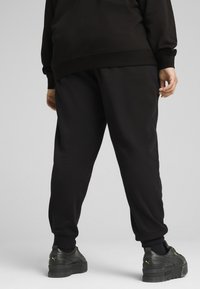 Schwarze Sweatpants mit elastischen Bündchen, aus Baumwollmaterial und entspanntem Schnitt. Kombiniert mit schwarzen Sneakers, die ein goldenes Logo-Detail aufweisen.