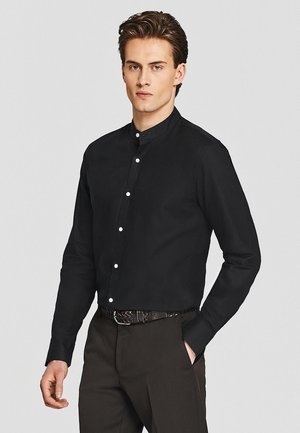 Mężczyzna noszący czarną koszulę z kołnierzem mandarynkowym i białymi guzikami, ciemne spodnie oraz pleciony pasek, stojący z jedną ręką w kieszeni.