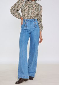 Ljust blå högmidjade jeans med vida ben, som har två framsidor och en texturerad yta, kombinerat med en blommig blus med långa ärmar.