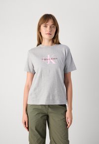 Camiseta de algodón gris con mangas cortas que presenta un logo en rosa y rojo de "Calvin Klein" en el centro, de corte casual y cuello redondo.