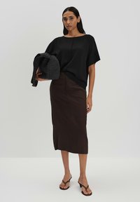 Maglietta nera oversize a maniche corte, gonna marrone lunga fino al ginocchio e sandali con tacco a strappo marroni. Tiene una borsa nera texture. Design minimal.