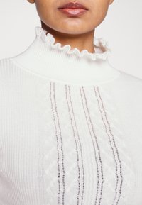 Der weiße, gerippte Rollkragenpullover verfügt über einen rüschengeschmückten Kragen, vertikale Muster und offene Strickdetails an der Vorderseite. Weiche Textur.