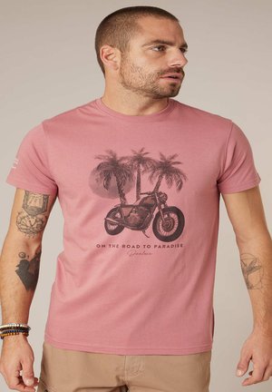 Homme portant un t-shirt rose avec une moto, des palmiers et le texte « Sur la route du paradis » imprimés sur le devant, un pantalon beige et des tatouages.