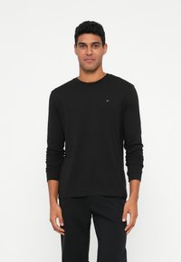 Homem vestido com uma camisa preta de manga longa simples e calças pretas, de pé contra um fundo branco.