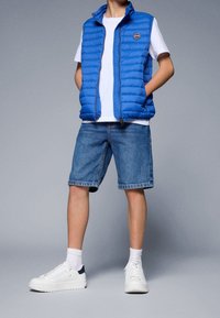 Blå vadderad väst med horisontell sömnad, vit T-shirt, jeansshorts och vita sneakers med marinblå detaljer. Avslappnad outfit.